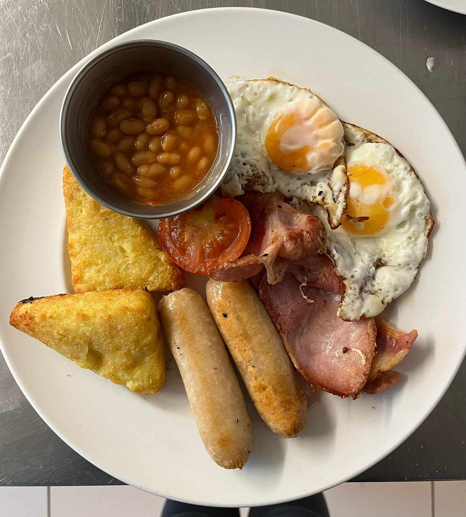 Full English breakfast with eggs, sausages, bacon, baked beans, and hash browns on a white plate.
