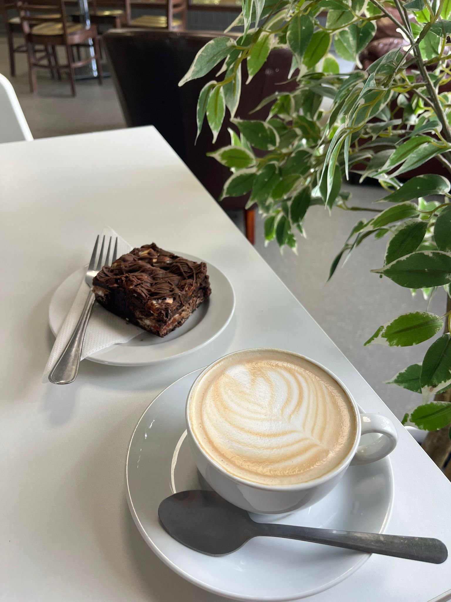 Cappuccino in a white cup with a latte art design, a piece of chocolate cake on a white plate, and a fork on a white table with a plant in the background.