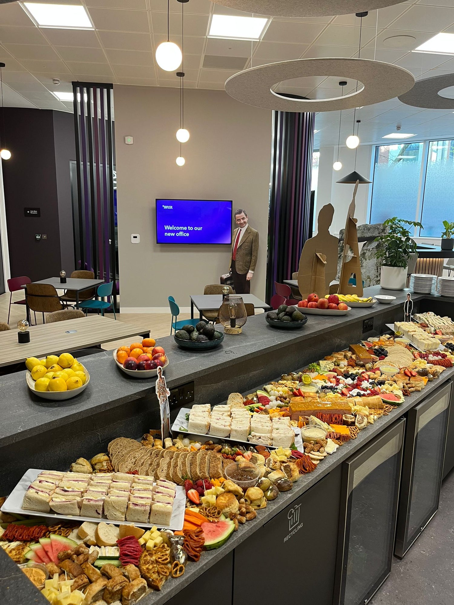 Buffet table with various food items in an office setting, featuring a TV screen on the wall.